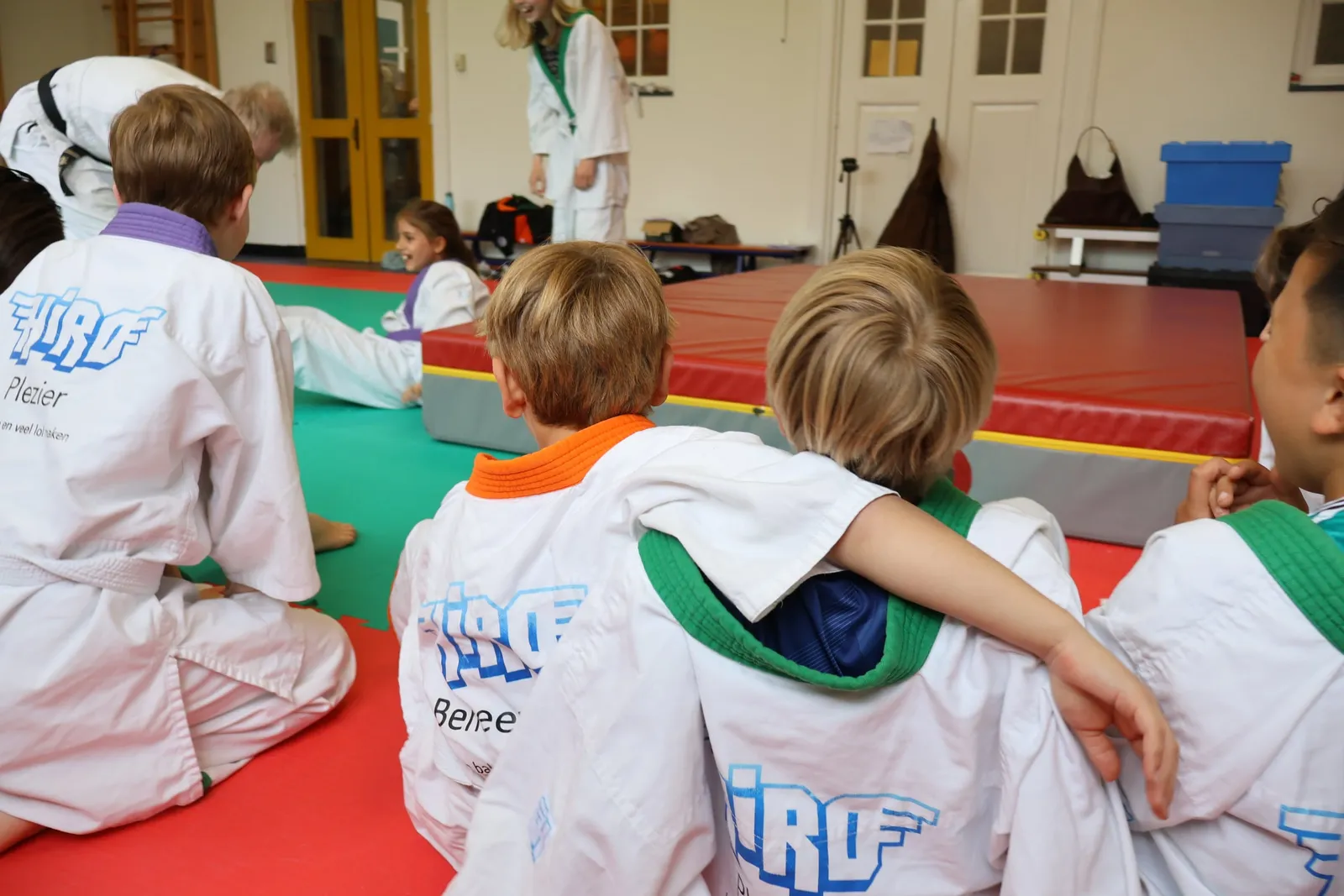 Twee kinderen zitten arm om schouder naast elkaar op de judomat